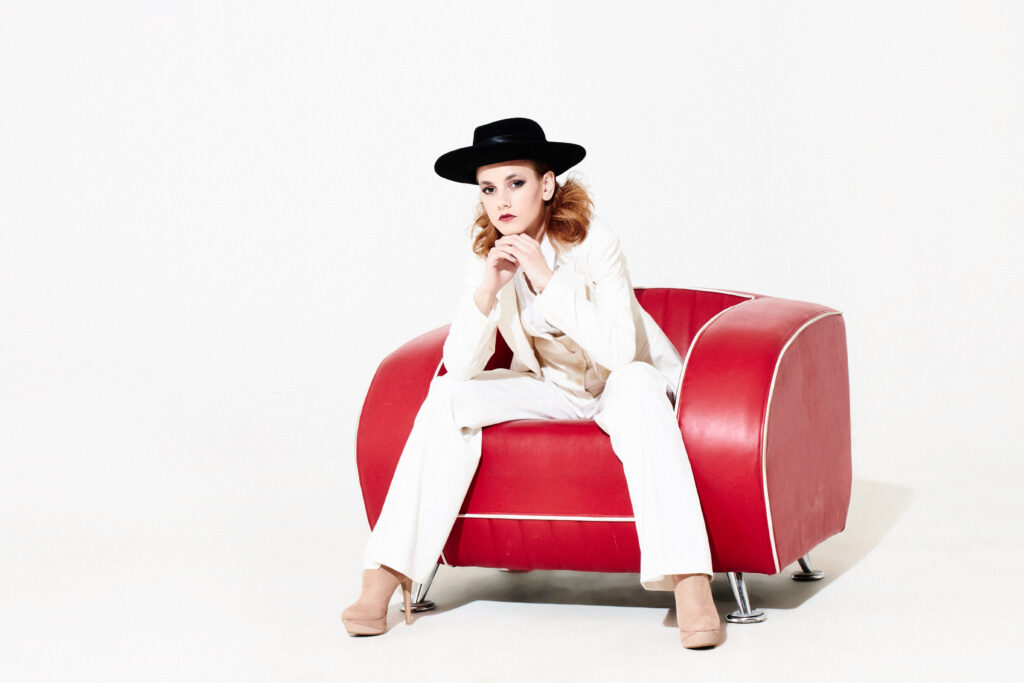 Fehér öltönyös, kalapos női modell piros műbőr fotelben ül.