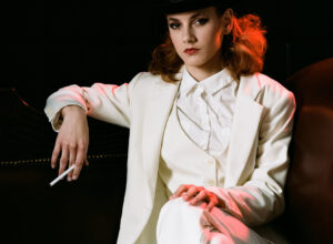 Öltönyös női modell kalapban, keresztbe tett lábbal cigarettázik
